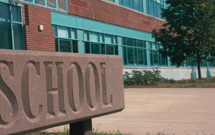 A stone sign with the word "SCHOOL" engraved on it stands in front of a brick school building with large windows and a tree nearby on a clear day.